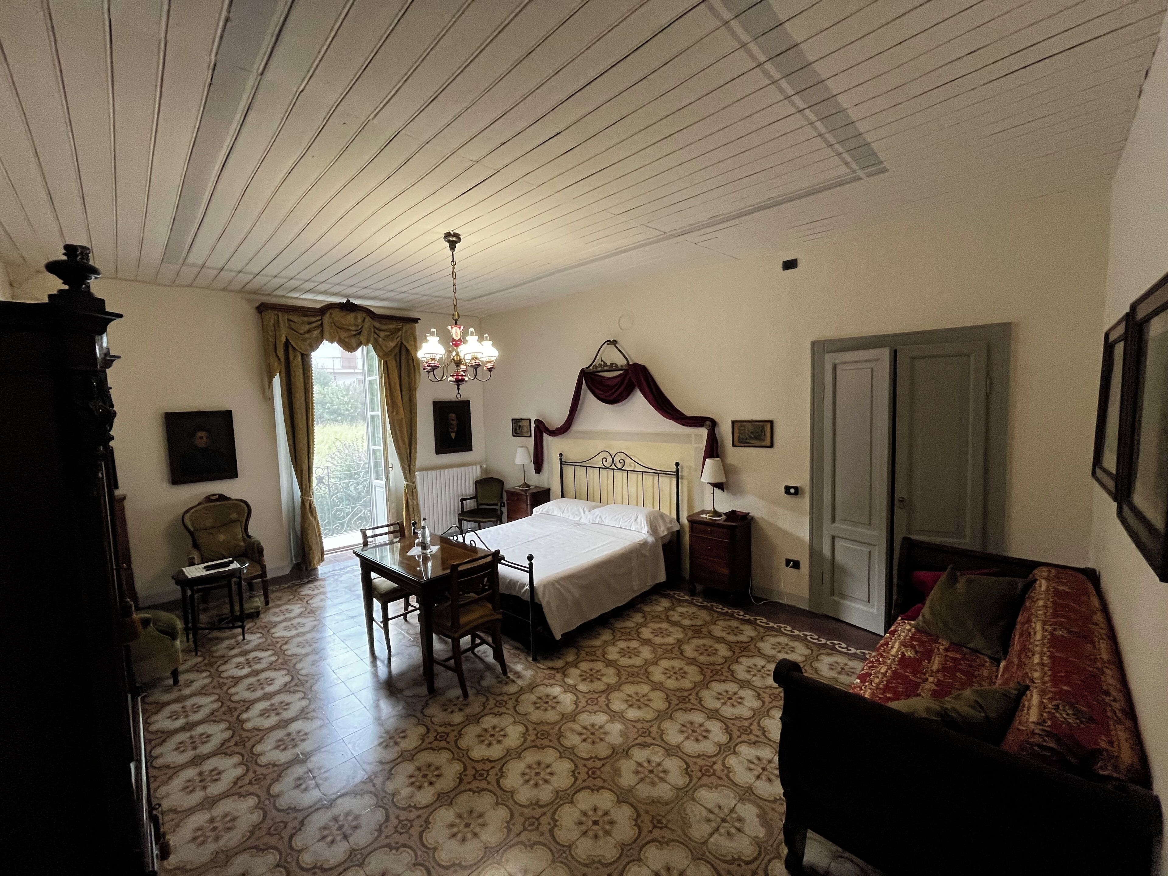 A bedroom with a bed, a chair, a table, a chandelier, and a door.