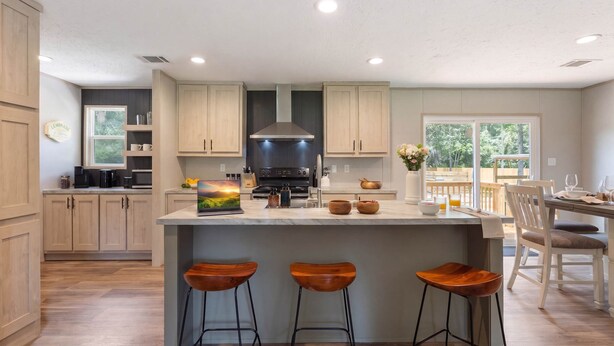 A modern kitchen with wooden cabinets, a central island with stools, a stove, and a dining area with chairs. - The Riverhead | Springs • Pavilion • Grill (Fort White)