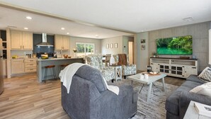 A modern living room with a grey sofa, a coffee table, and a TV. There is a kitchen area with wooden cabinets and a dining space with patterned chairs. - The Riverhead | Springs • Pavilion • Grill (Fort White)