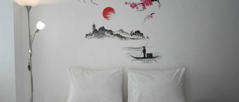 A bedroom with a bed, two pillows, a nightstand, and a wall with a painting of cherry blossoms, a sun, and a mountain scene.
