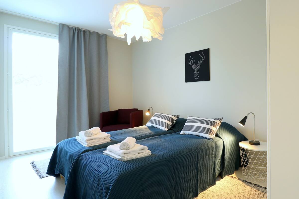 A modern bedroom with two beds, a large window with curtains, a wall-mounted artwork, and a round side table with a lamp.