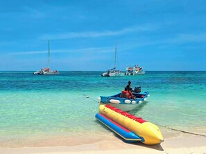 A beach with clear turquoise water, a small inflatable boat, and two larger boats in the distance.