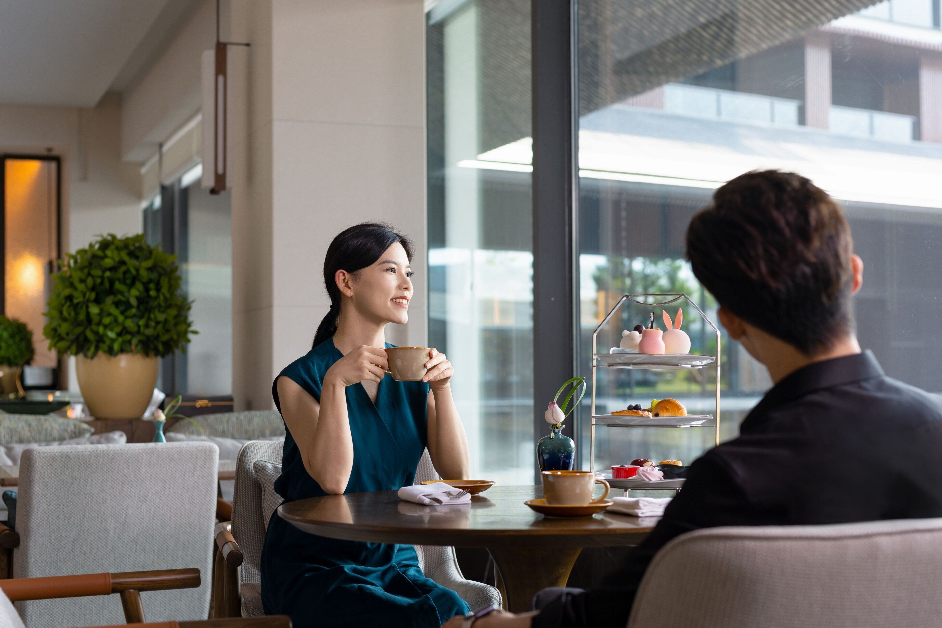 Uma mulher e um homem sentados à mesa em um café moderno com uma janela grande, uma planta e um serviço de chá.