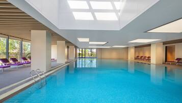 Una piscina interior con agua azul cristalina, rodeada de sillas reclinables moradas y un diseño de techo moderno.