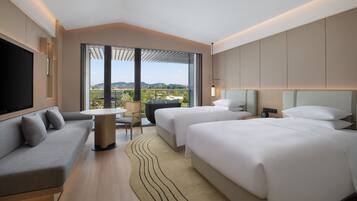 A modern hotel room with a sofa, two beds, a TV, a table, and a view of the city.