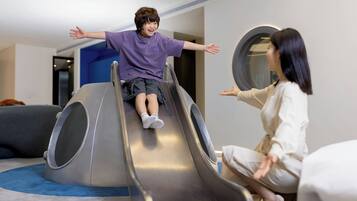 A child sliding down a metallic slide in a room with a blue carpet and a circular mirror on the wall.
