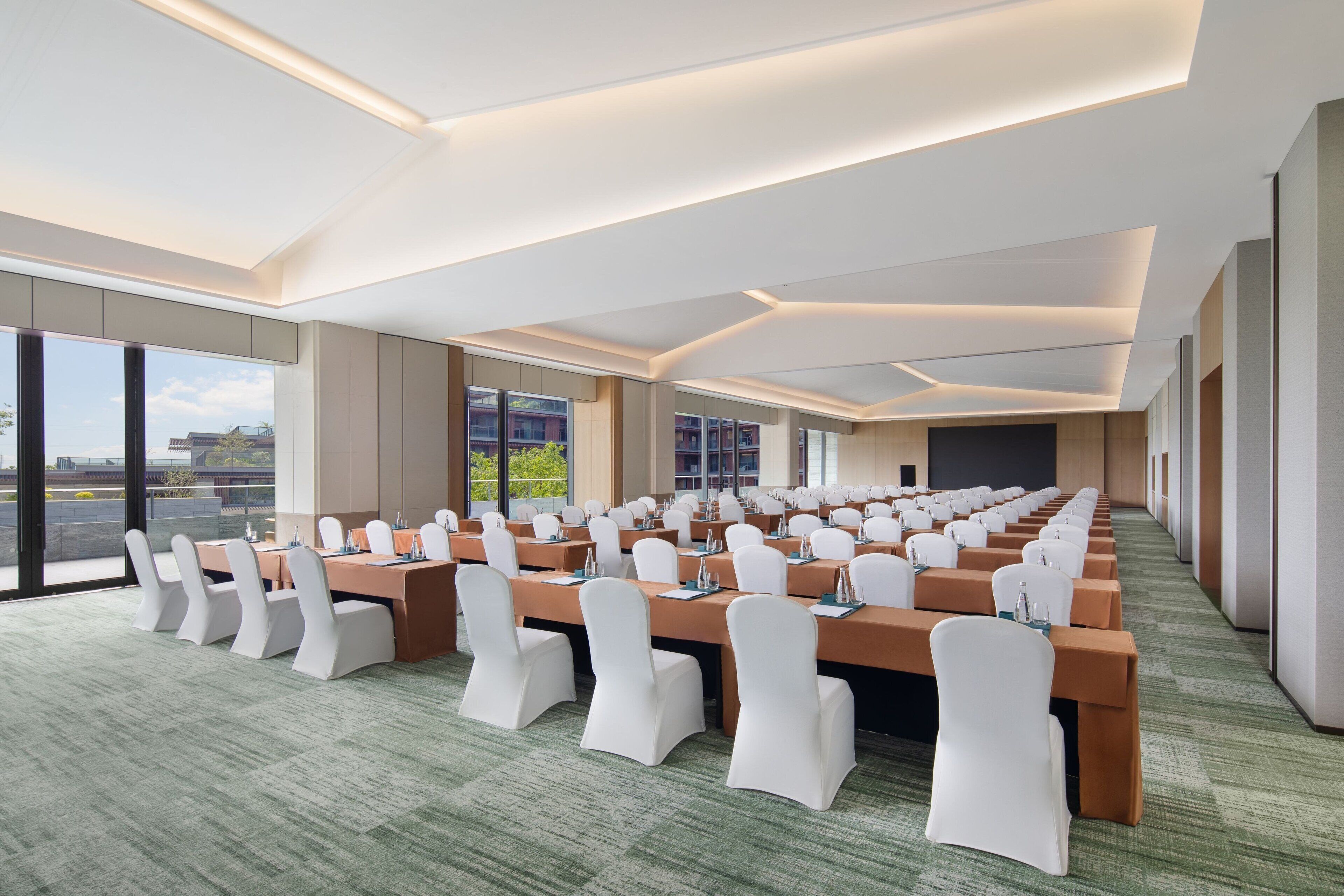 A conference room with rows of tables and chairs, large windows, and a green carpeted floor.