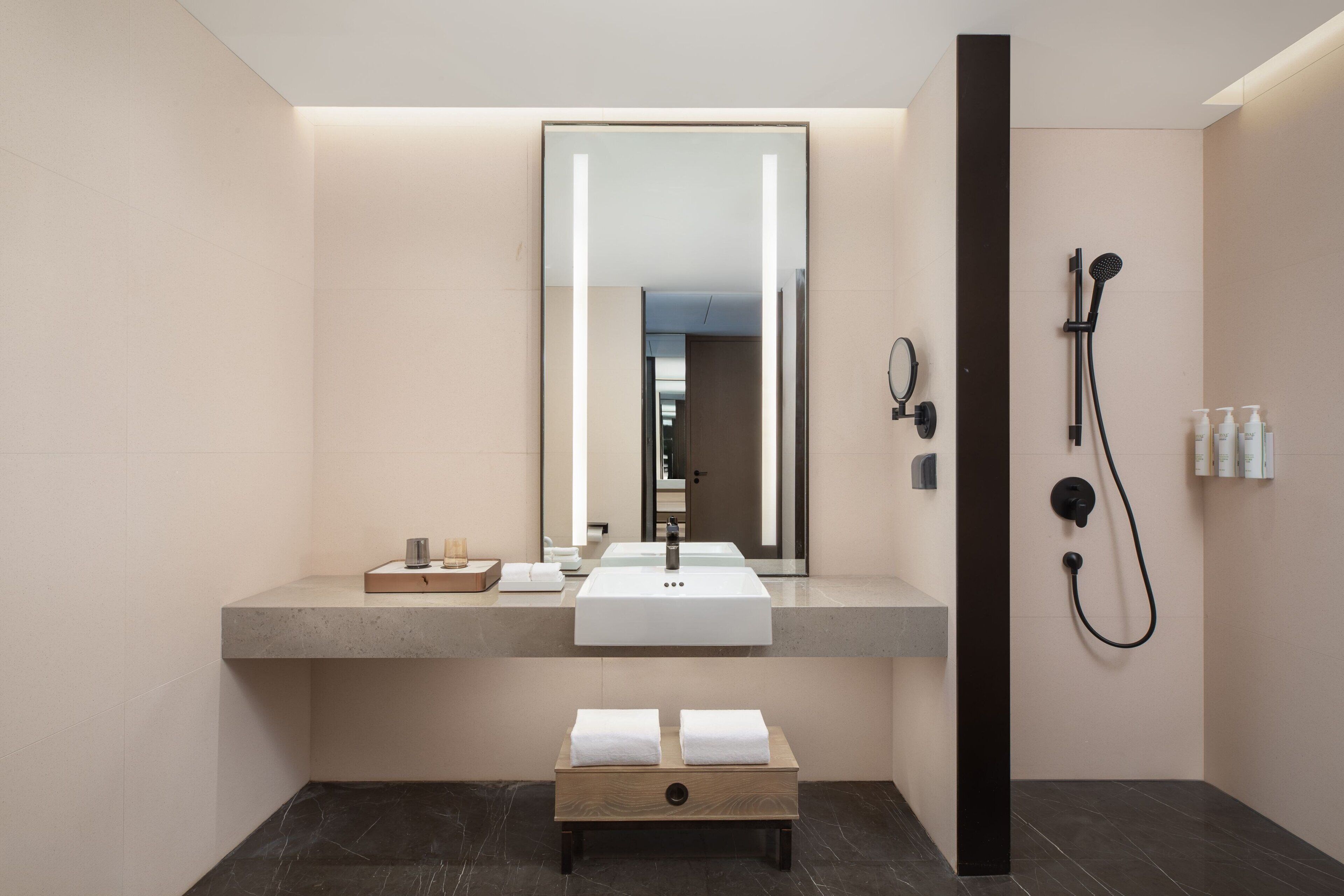 A modern bathroom with a large mirror, a sink, a shower, and a bench with towels.