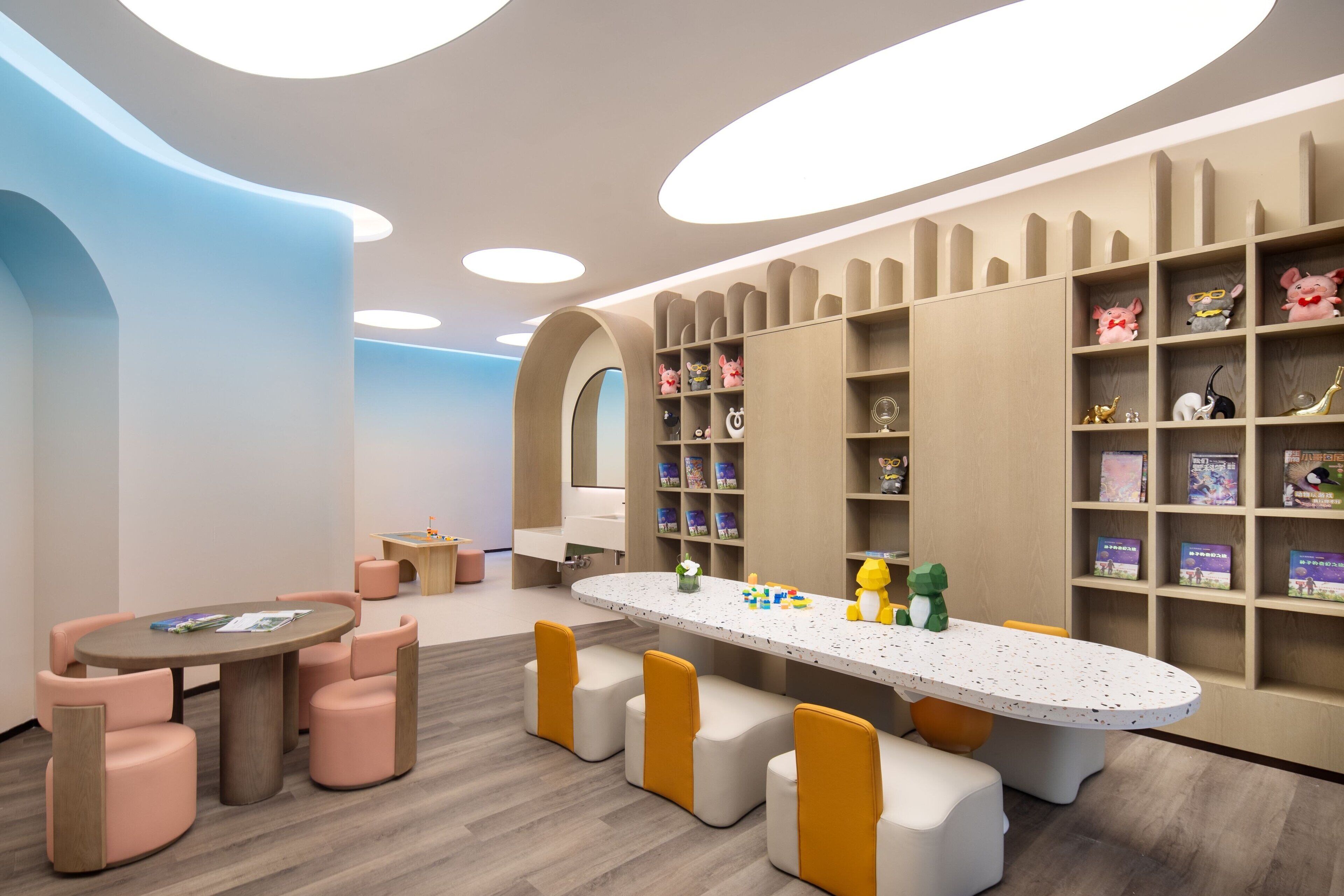 A children's play area with a long table, cushioned seating, and shelves with toys and books.