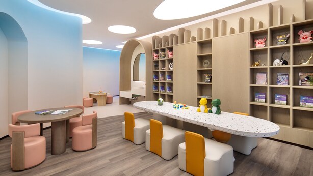 Un espace de jeux pour enfants avec une grande table, des sièges rembourrés et des étagères remplies de jouets et de livres.