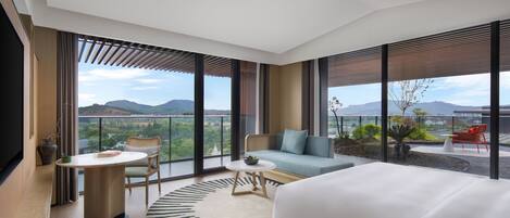 A modern hotel room with a large bed, a round coffee table, a sofa, and a dining table. The room has large windows offering a view of the mountains and a balcony with red chairs.