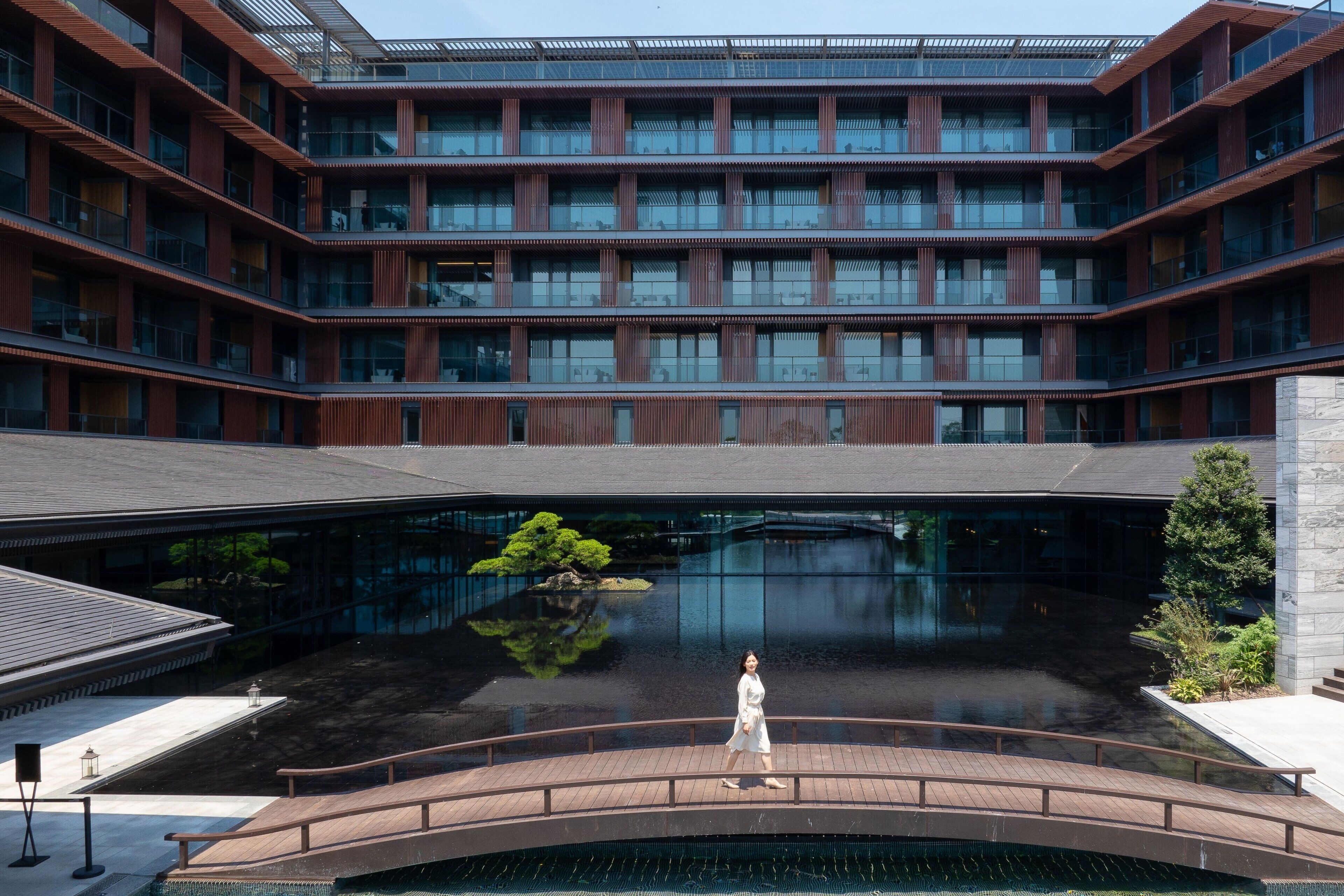 A modern building with multiple floors, a central courtyard with a reflective water feature, and a bridge with a person walking.