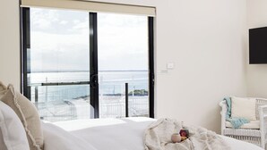 A bedroom with a large window overlooking a balcony with a railing. There is a bed with a white comforter and pillows, a small table with a cup and a plate of cookies, and a white wicker chair with a cushion.