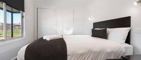 A modern hotel room with a large bed, a dark headboard, a bedside table, and a window with a view of a building and greenery.
