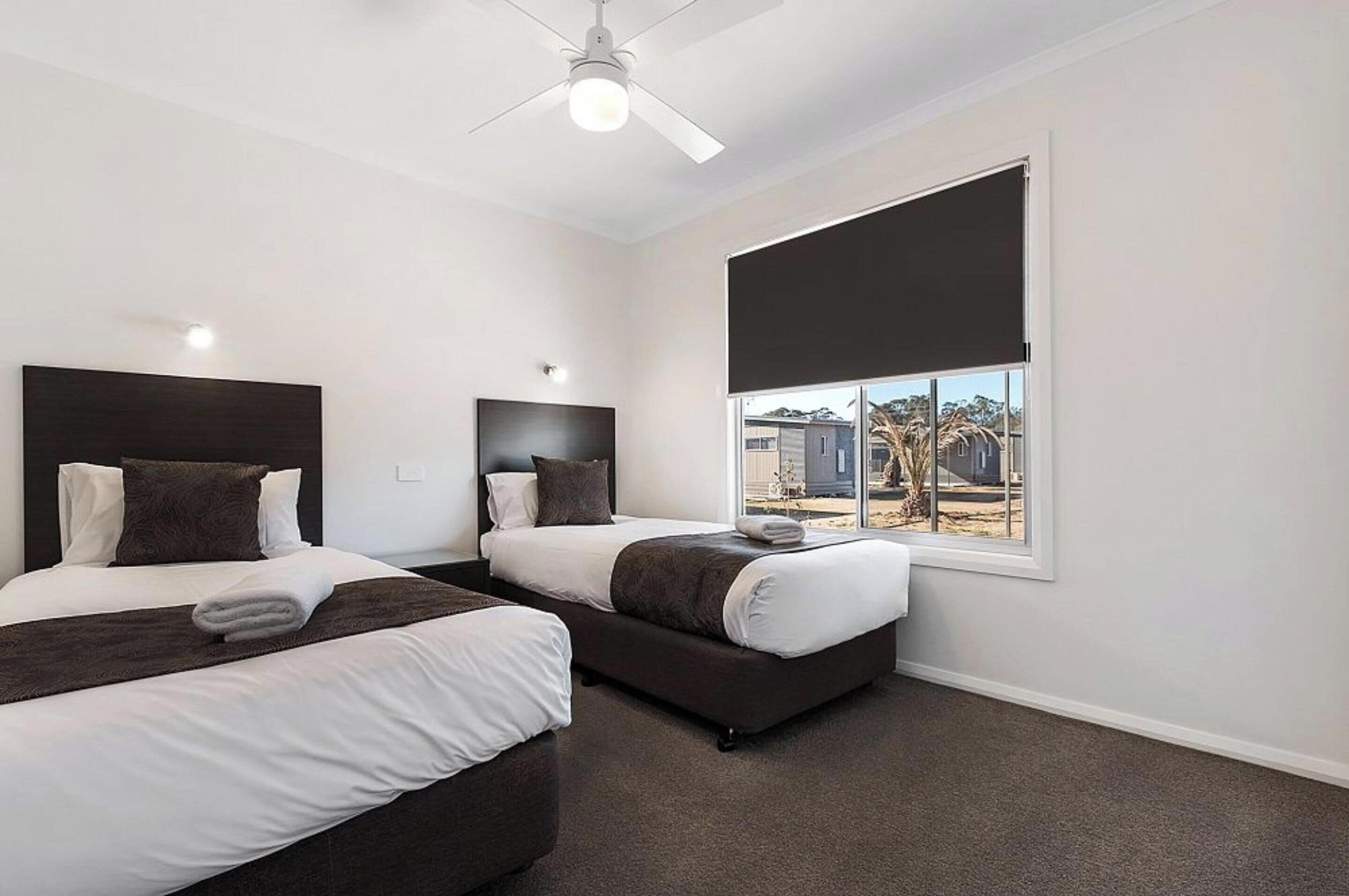 A hotel room with two beds, a ceiling fan, a large window with a view of the outside, and a dark-colored carpet.