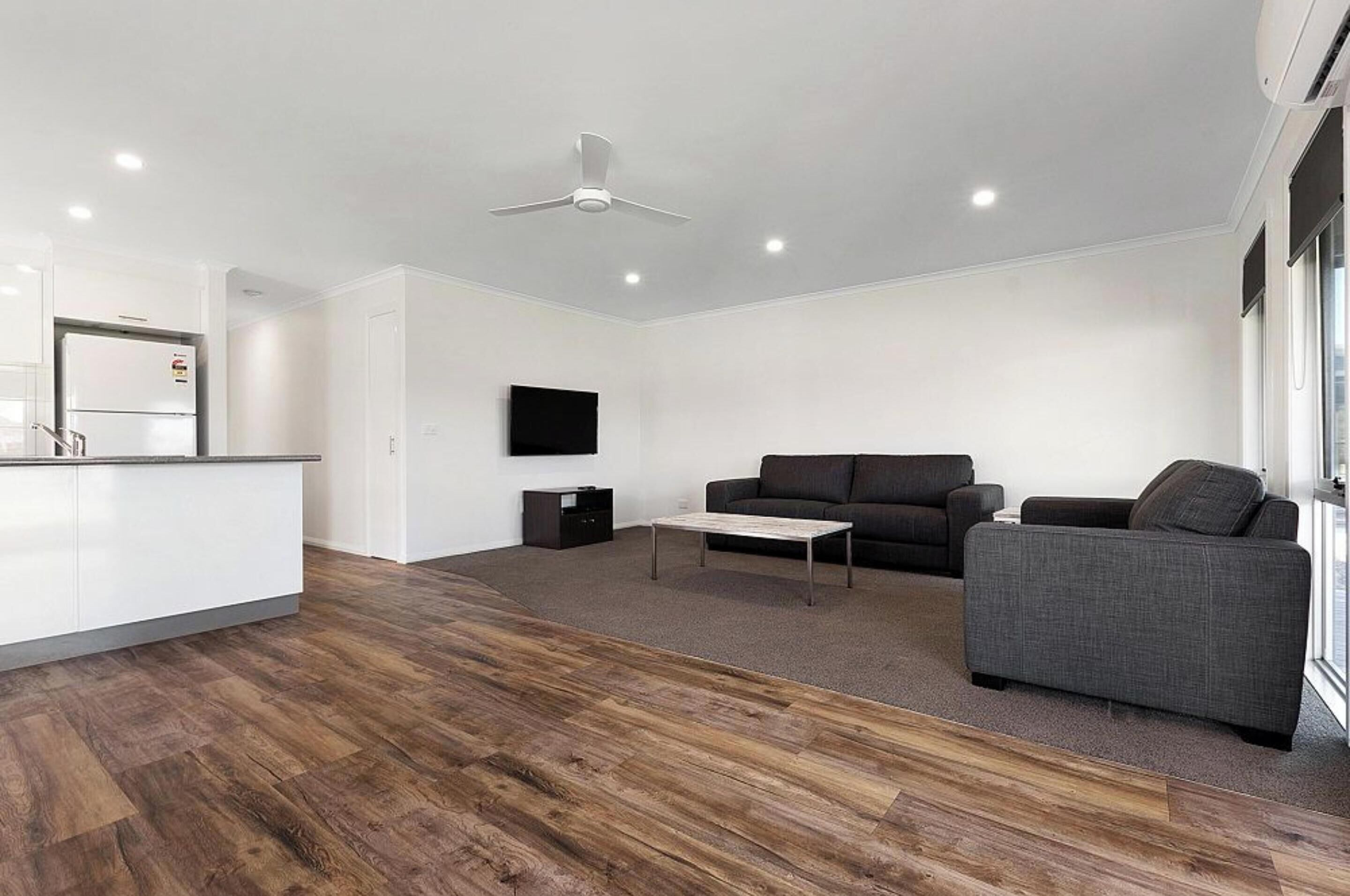 Een moderne woonkamer met een houten vloer, een grijze bank, een salontafel, een flatscreen-televisie aan de wand en een plafondventilator.