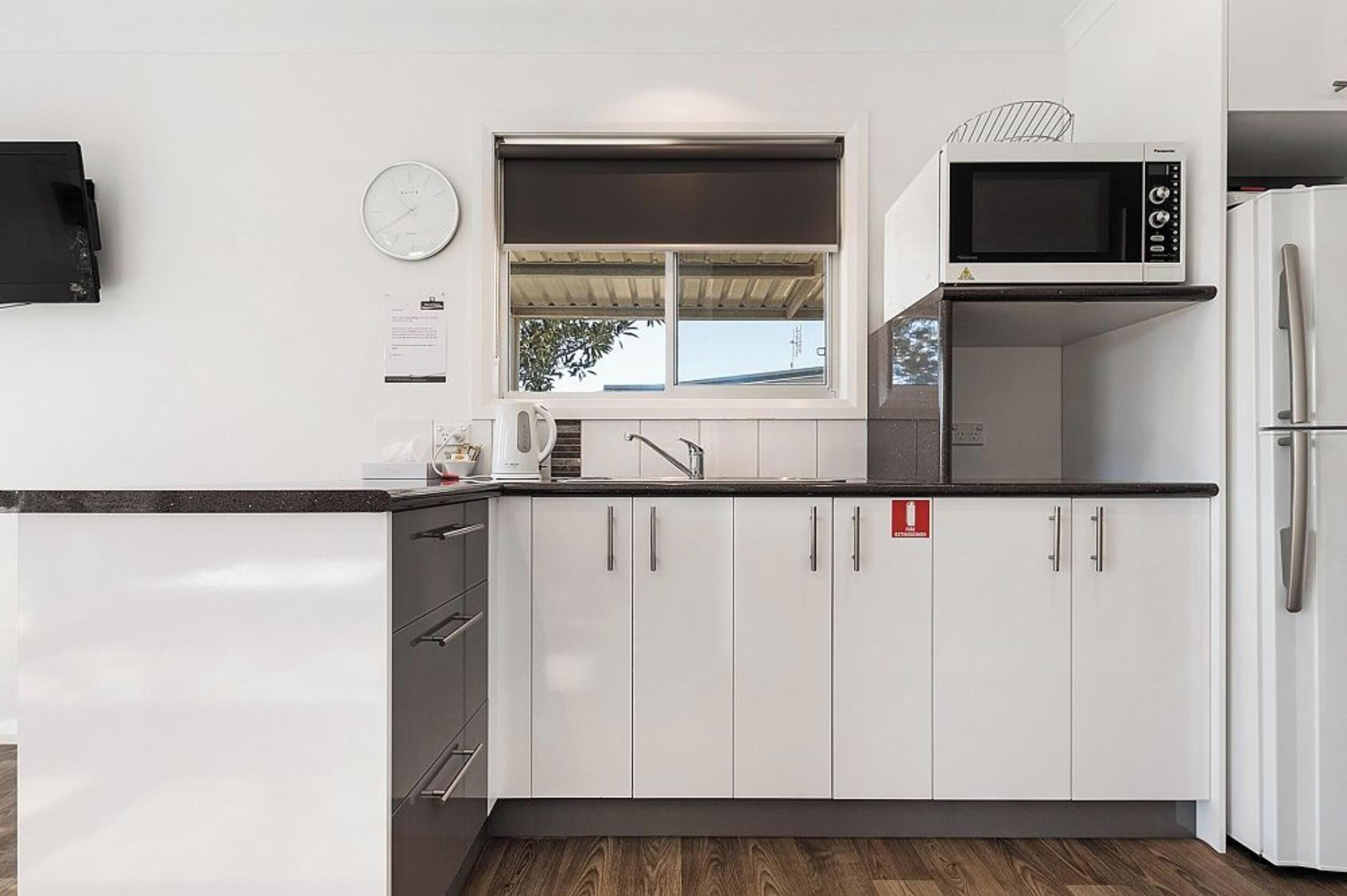Una cocina moderna con armarios blancos, encimera negra, microondas, refrigerador, reloj y ventana con vista al exterior.