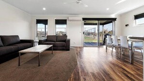 A modern living room with a sofa, coffee table, and dining area with chairs. There is a ceiling fan, an air conditioning unit, and a view through the glass door. - Discovery Parks Moama Waters  (Moama)