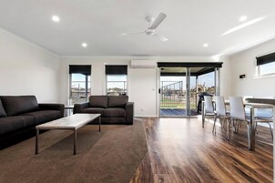 Una sala de estar moderna con sofá, mesa de centro y comedor con sillas. Hay un ventilador de techo, un equipo de aire acondicionado y vistas a través de la puerta de cristal.