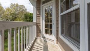 Un porche couvert avec une balustrade blanche, une porte dotée d’une fenêtre et une vue sur les arbres et la verdure.