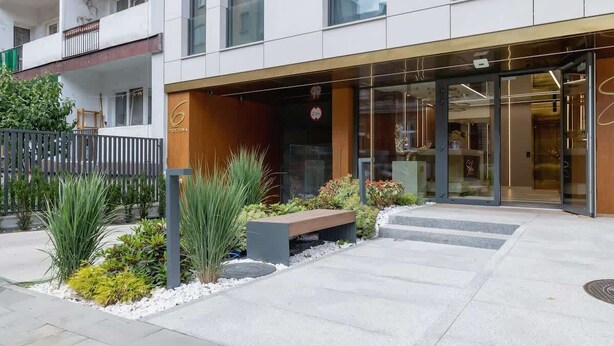 Un edificio moderno con entrada de vidrio, un banco de madera y un jardín ajardinado.