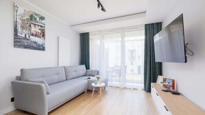 Una sala moderna con un sofá gris, una mesita redonda con un florero, un televisor de pantalla plana montado en la pared y una ventana con cortinas.