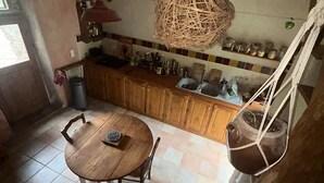 A rustic kitchen with wooden cabinets, a round wooden table, and a hanging wicker light fixture. - Self-contained, eco-renovated farmhouse in a small, unspoilt valley in the Cévennes. (Gagnières)