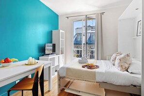 Ein kleines Zimmer mit einem Bett, einem Schreibtisch mit Stuhl, einem Fernseher und einem Fenster mit Blick auf Gebäude.