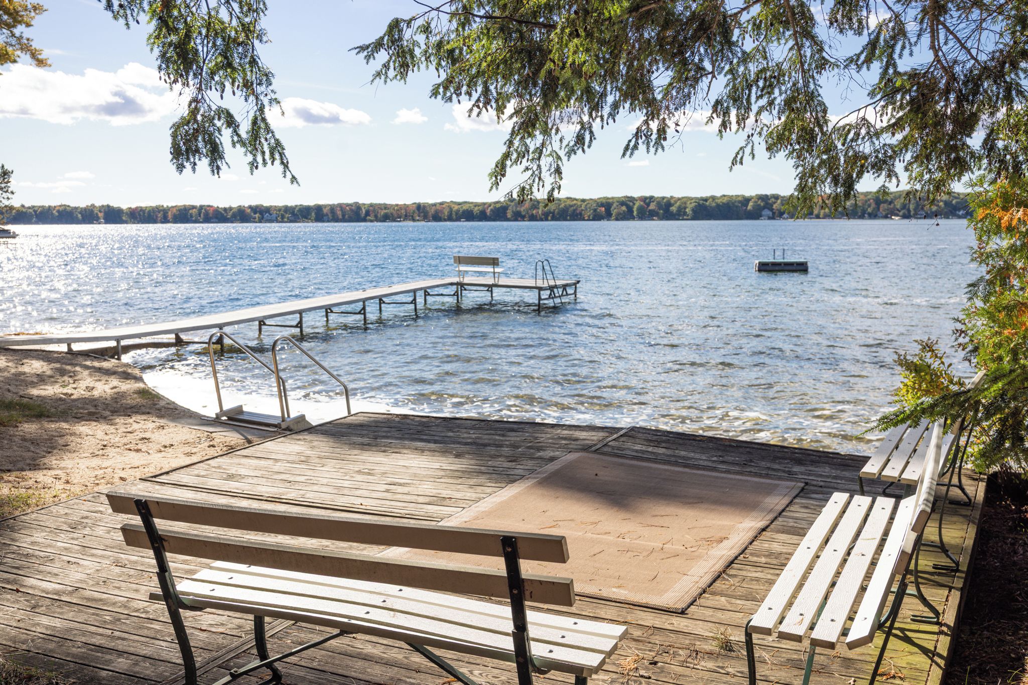 A wooden deck with benches overlooking a calm lake, a dock extending into the water, and a distant boat.