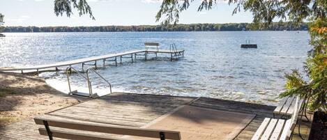 A wooden deck with benches overlooking a calm lake, a dock extending into the water, and a distant boat.