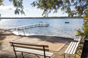 Eine Holzterrasse mit Bänken bietet Blick auf einen ruhigen See, eine Anlegestelle erstreckt sich ins Wasser, und ein Boot ist in der Ferne sichtbar.