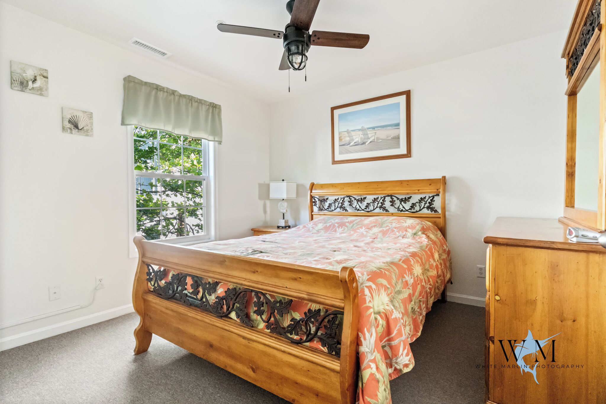 A bedroom with a wooden bed, a ceiling fan, a window with a view of greenery, a bedside table with a lamp, and a wooden dresser.