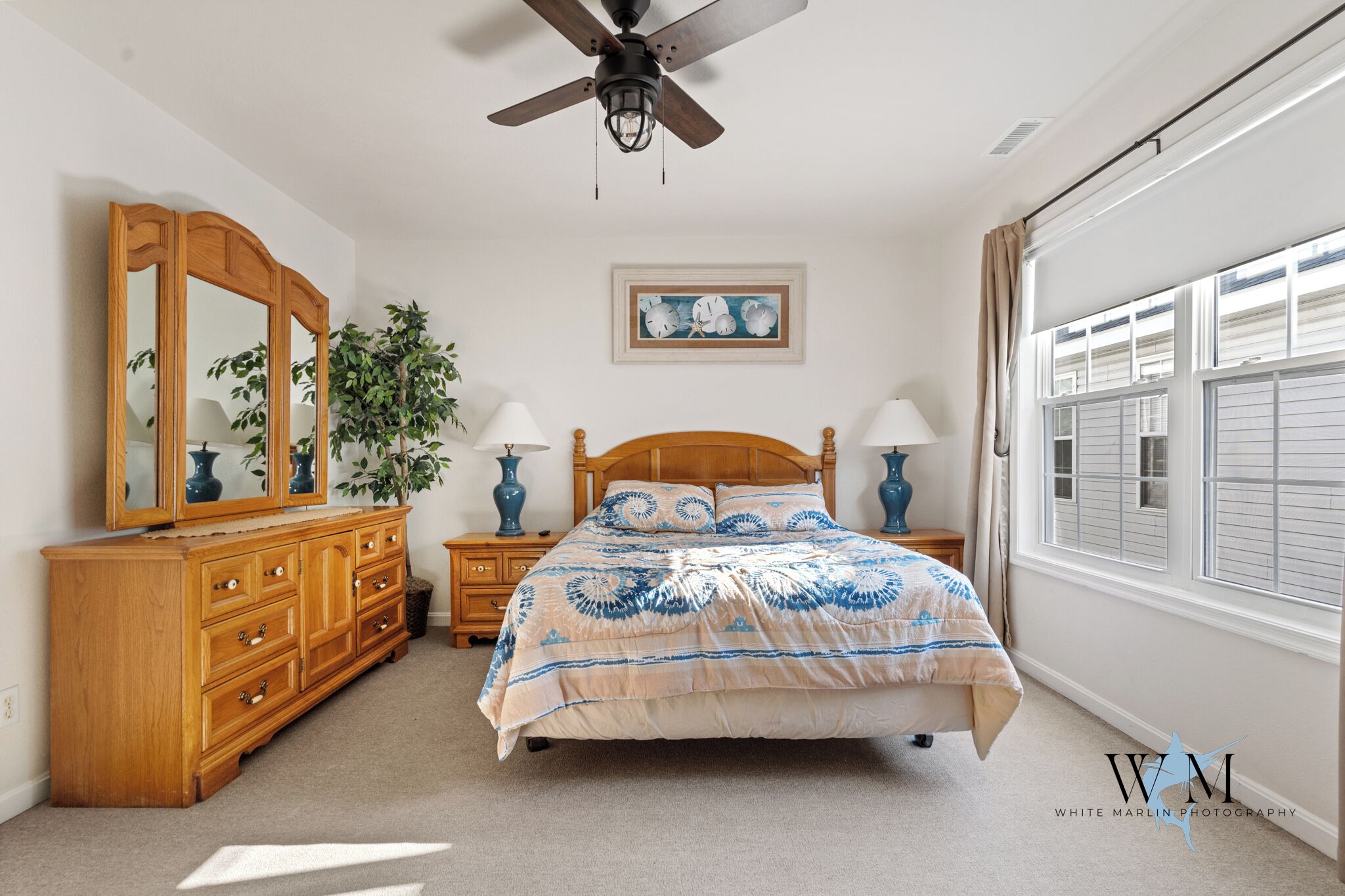 A bedroom with a bed, wooden dresser, two nightstands with lamps, a ceiling fan, a window with blinds, and a framed picture on the wall.