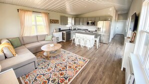 Un salón amplio con sofá, mesa de centro y alfombra estampada. Se ve una cocina con electrodomésticos modernos y una zona de comedor con taburetes de bar.