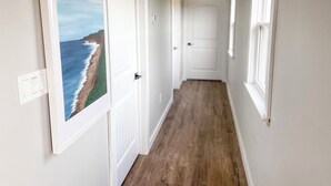 Un pasillo con piso de madera, puertas blancas y un cuadro con un paisaje costero.