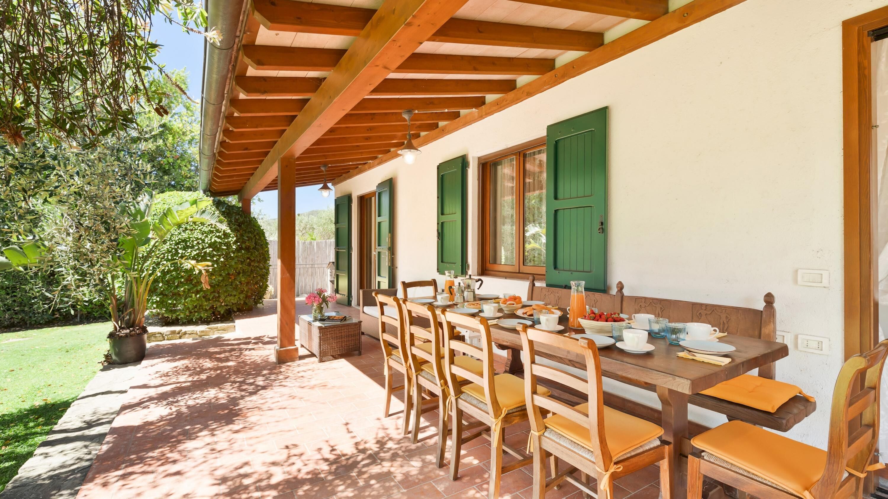 An outdoor dining area with a wooden table and chairs, green shutters, and a potted plant.