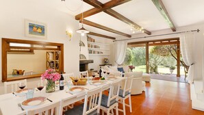 A dining area with a white table set for four, surrounded by white chairs. There is a bookshelf with books and decorative items, a fireplace, and a view of the garden through large windows.