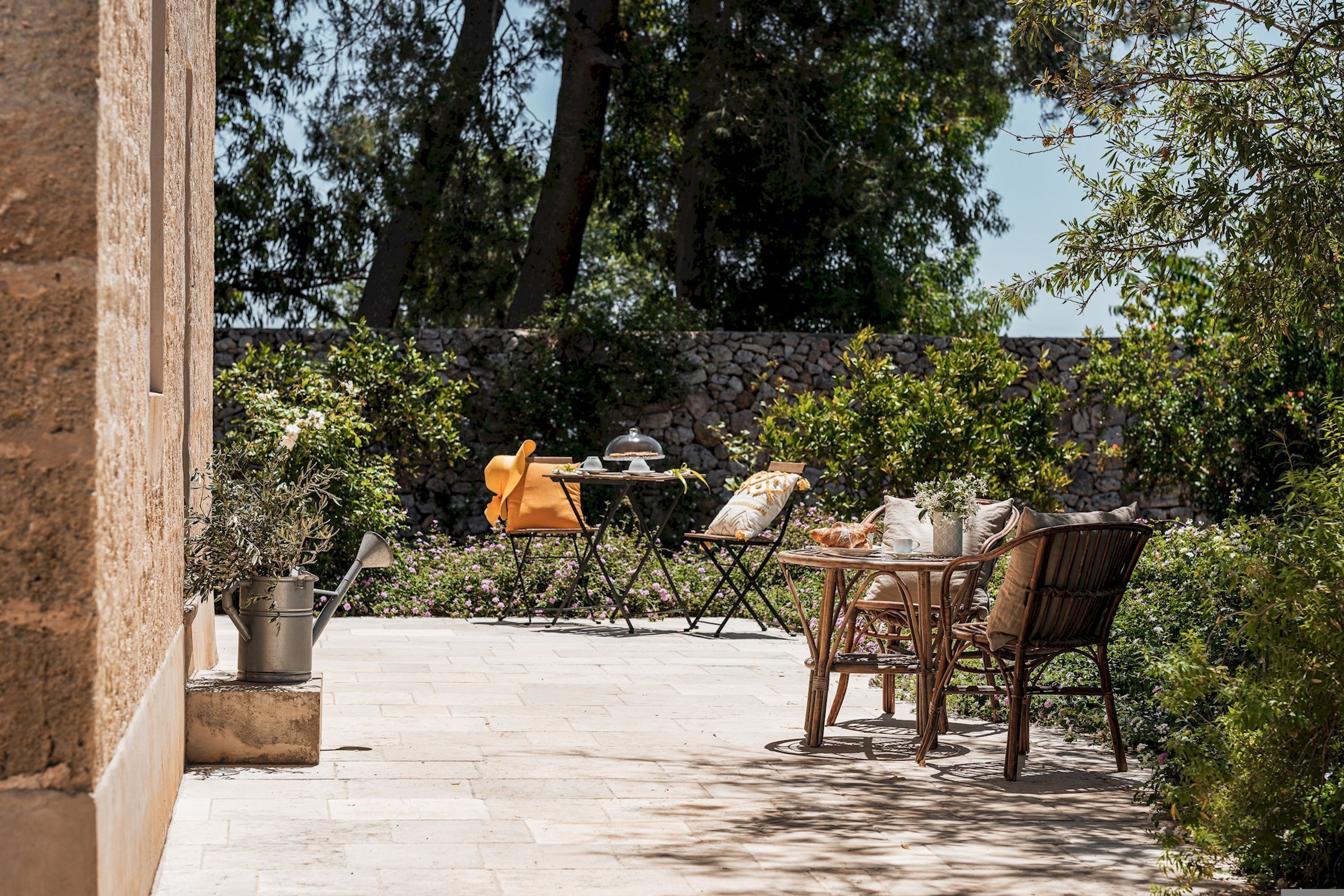 Um pátio externo com mesa e cadeiras, um vaso de planta e um muro de pedra ao fundo.