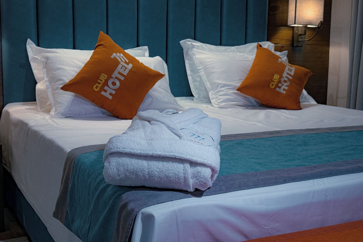 A hotel room with a bed, two pillows with 'Club Hotel' branding, a white towel, and a nightstand with a lamp.