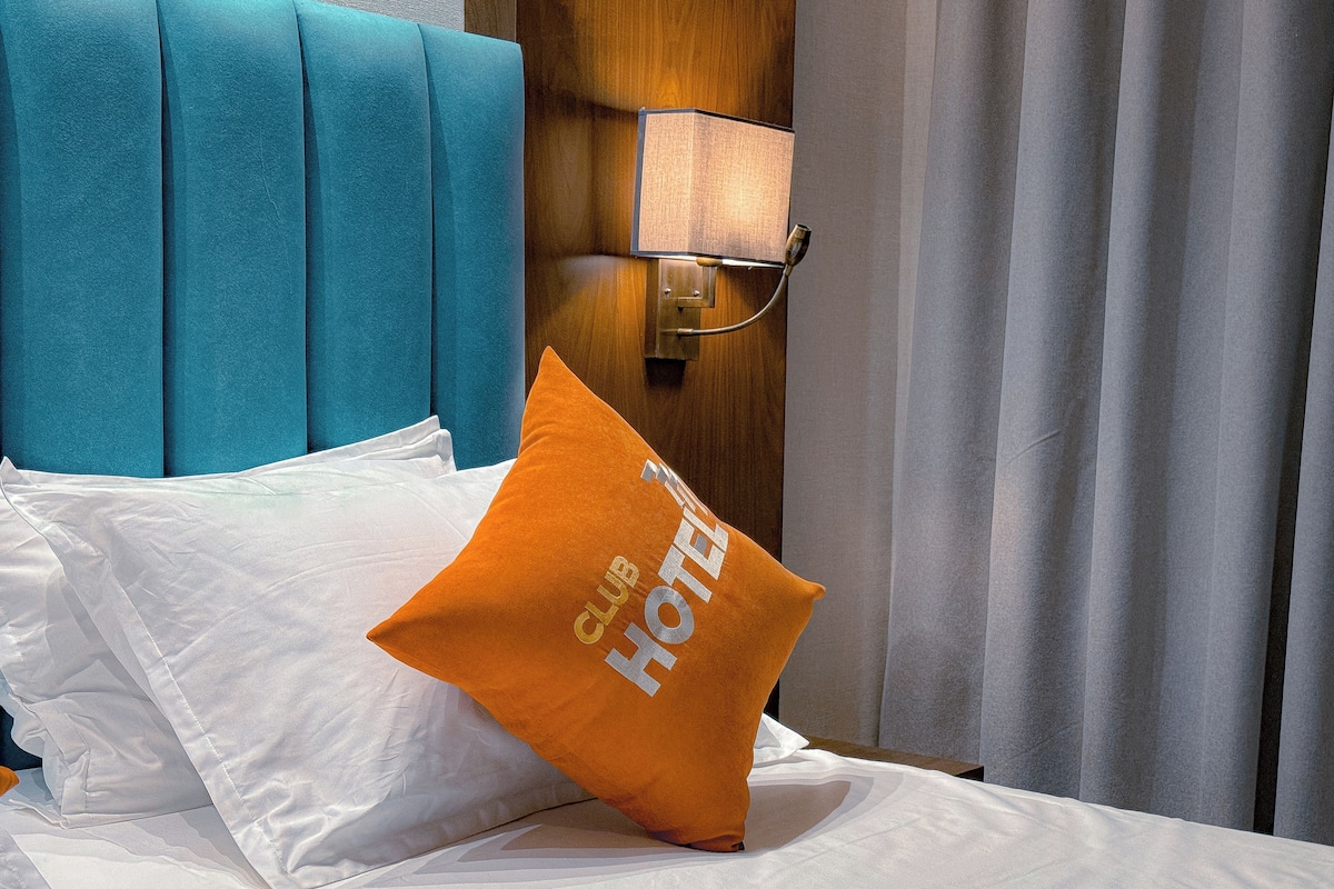 A hotel room with a teal headboard, a white bedspread, and an orange pillow with "CLUB HOTEL" text.