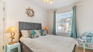Une chambre à coucher comprenant un lit, deux lampes de chevet, une décoration murale représentant un gouvernail de bateau, une fenêtre avec des rideaux et un fauteuil.