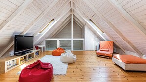 Une chambre mansardée avec un plafond en pente, des poutres apparentes, une télévision, des fauteuils en forme de poire et un canapé.