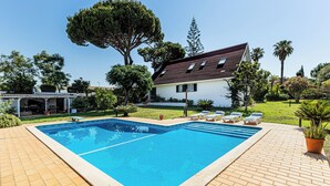 Une piscine rectangulaire bordée d’une terrasse carrelée, entourée d’une maison blanche au toit foncé, d’arbres et d’un ciel bleu clair.