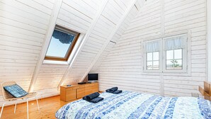 Une chambre avec un plafond en pente, un lit recouvert d’une couvre-lit à motifs floraux, une commode en bois, un téléviseur et une fenêtre donnant sur des palmiers.
