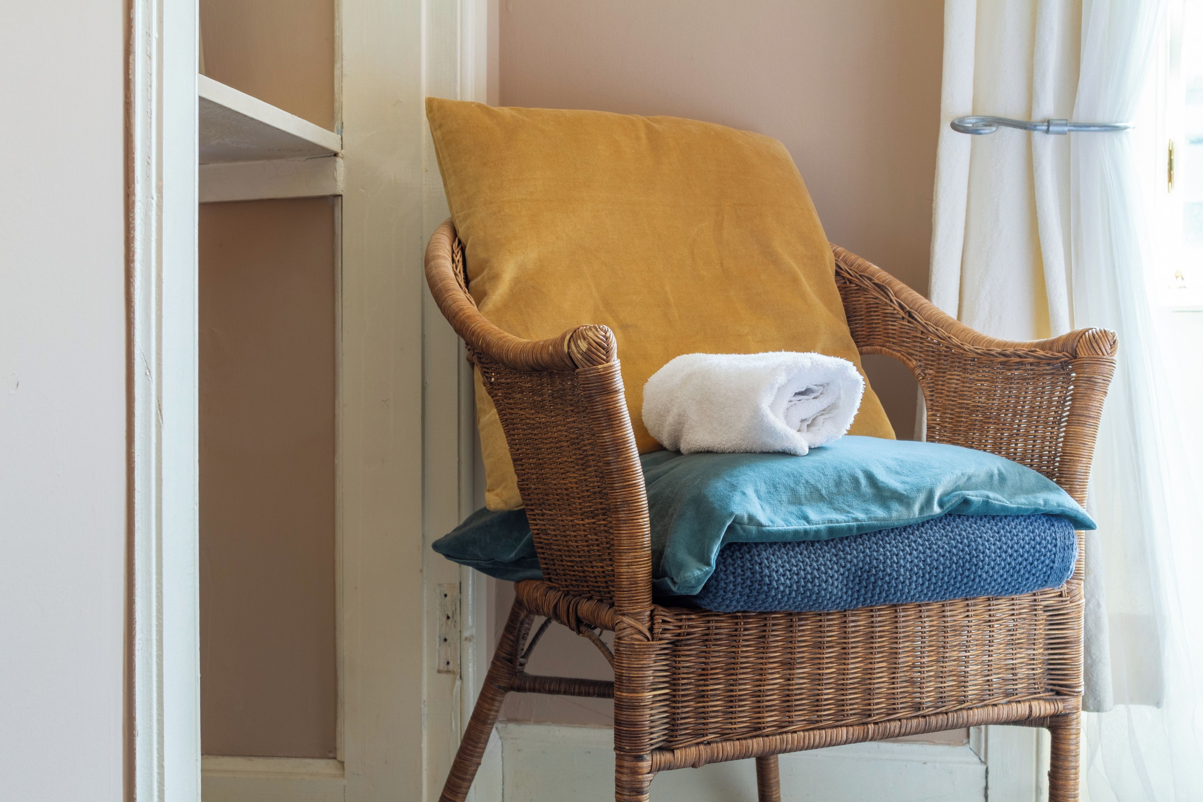 A wicker chair with a yellow pillow and a folded white towel, positioned near a window with sheer curtains.