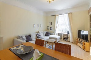 Ein modernes Wohnzimmer mit einer Couch, einem Esstisch, einem Fernseher und einem Fenster mit Vorhängen.