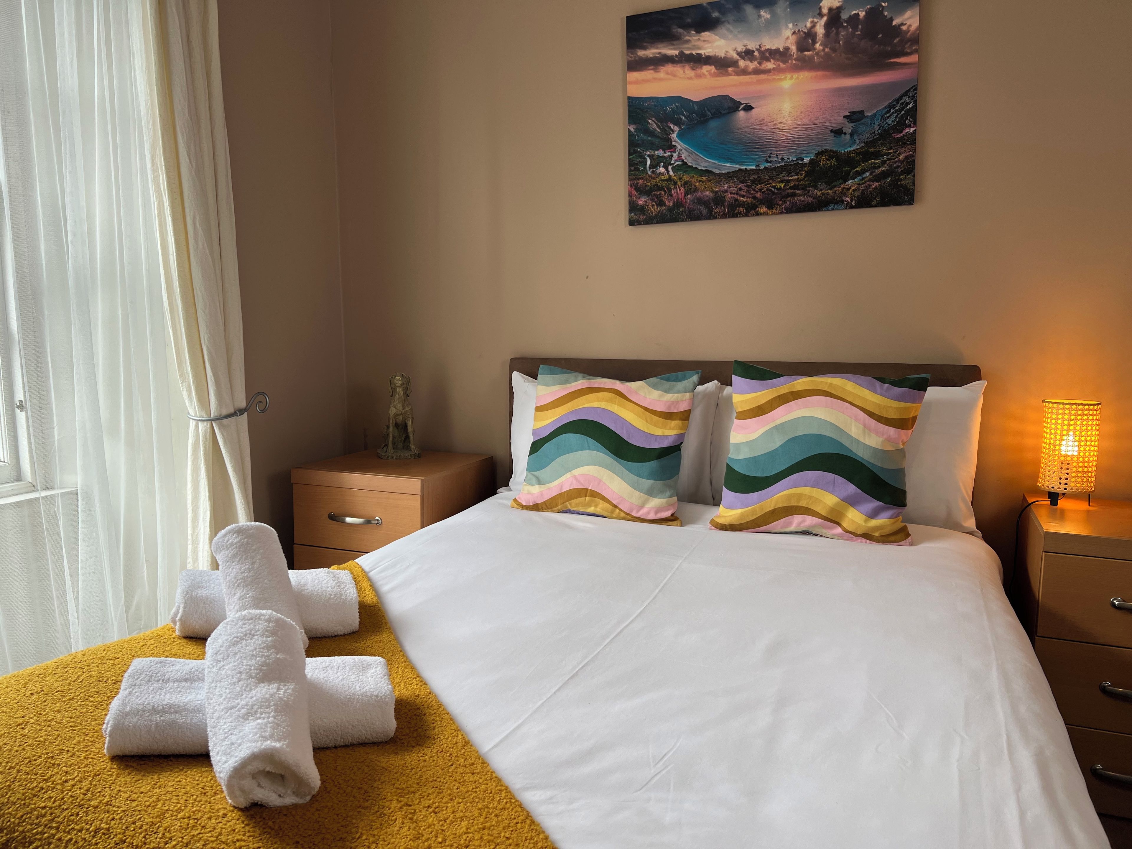 A hotel room with a bed, two pillows, a nightstand, a lamp, a painting, and a window with curtains.