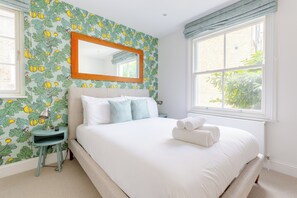A bedroom with a large bed, a bedside table, a mirror, and a window with a view of greenery.