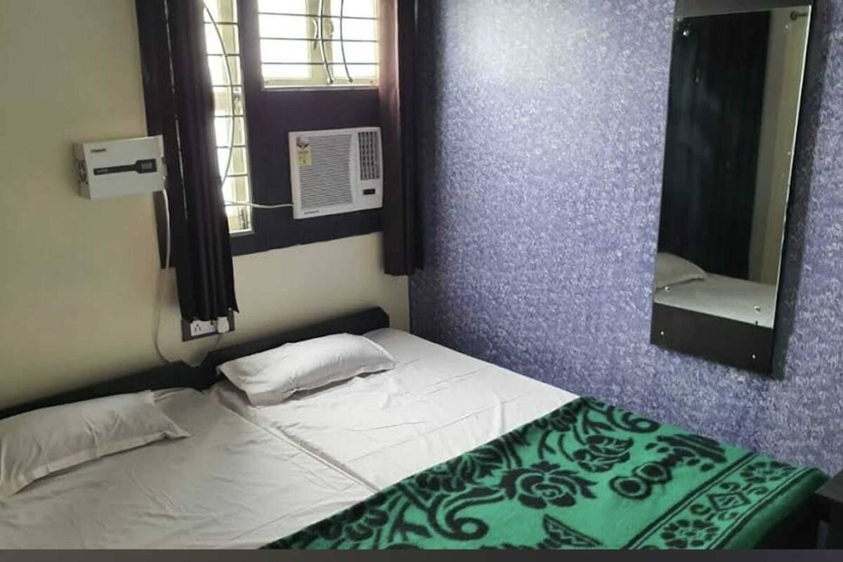A single bed with white linens, a green patterned blanket, a wall-mounted air conditioner, and a mirror.