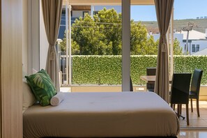 A bedroom with a bed, a green pillow, a view of a hedge and buildings, and a balcony with a table and chairs. - Sweet Stay (Cape Town)